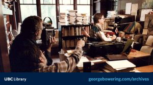 A photo of George Bowering in his creative work space, seated at his desk to the right, with a cameraperson filming to the left